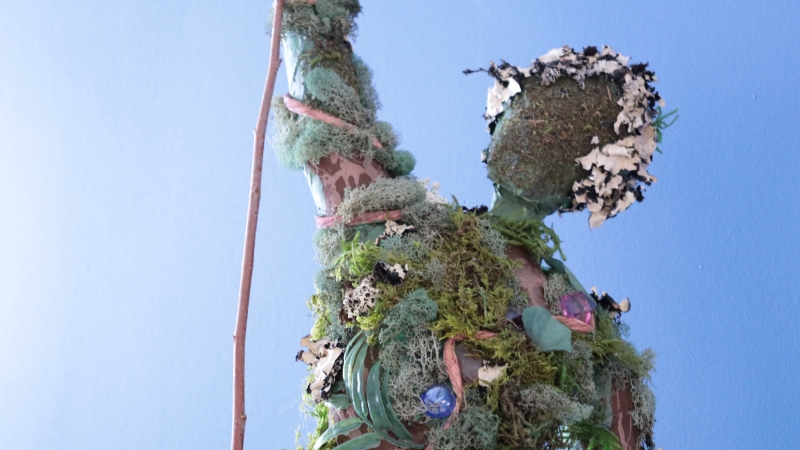 Student sculpture of a human figure covered in moss, lichen, and natural greenery against a blue background.