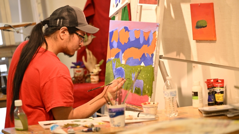 Student wearing a baseball cap painting a landscape with animals on an easel.
