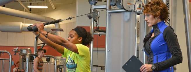 A woman in a neon shirt uses a cable weight machine while a trainer with a clipboard guides her in the fitness center.